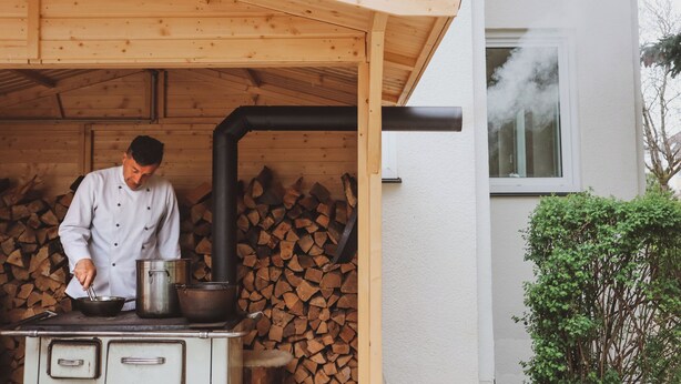 Un cocinero preparando comida al aire libre junto a una estructura de madera, una estufa y una pila de leña.