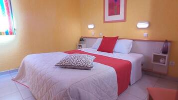 Une chambre d’hôtel avec un lit, des tables de chevet, une petite table, une couvre-lit rouge et blanche, un coussin à motifs et un tableau au mur.