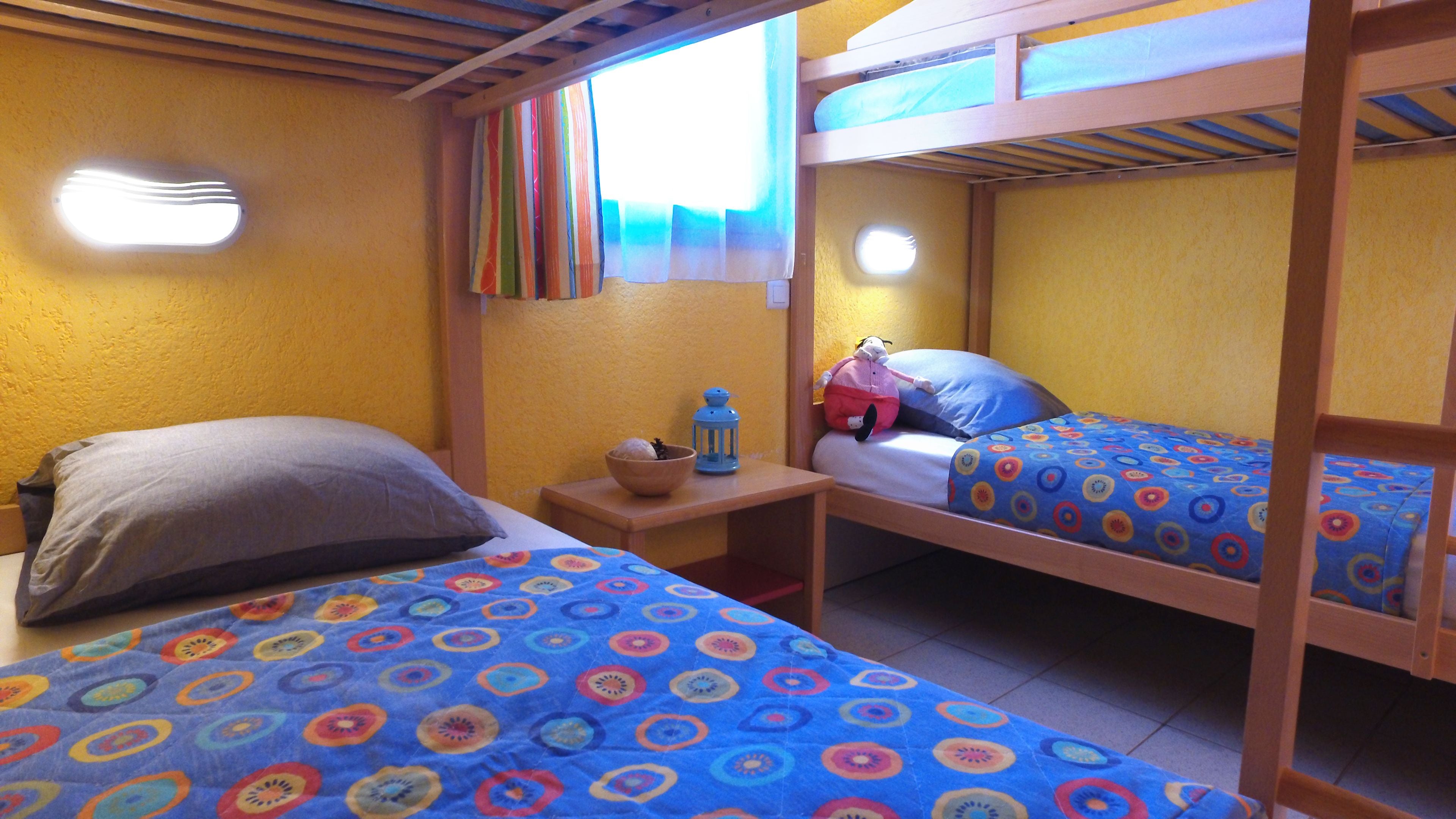 A room with two bunk beds, a small table with a bowl and a lantern, and a window with colorful curtains.