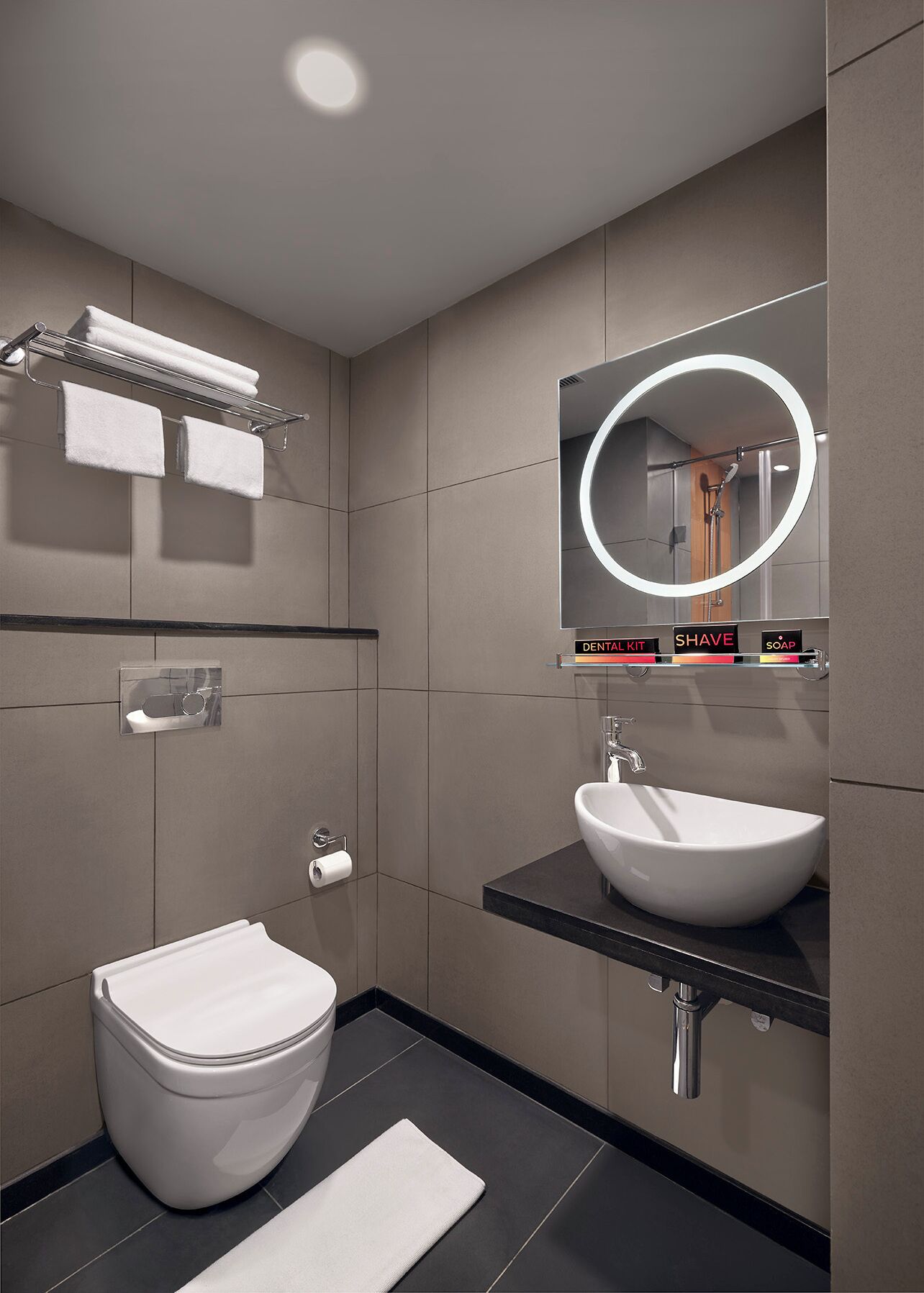 A modern bathroom with a toilet, sink, and a mirror with a built-in light. There is a towel rack above the toilet and a shelf with toiletries.