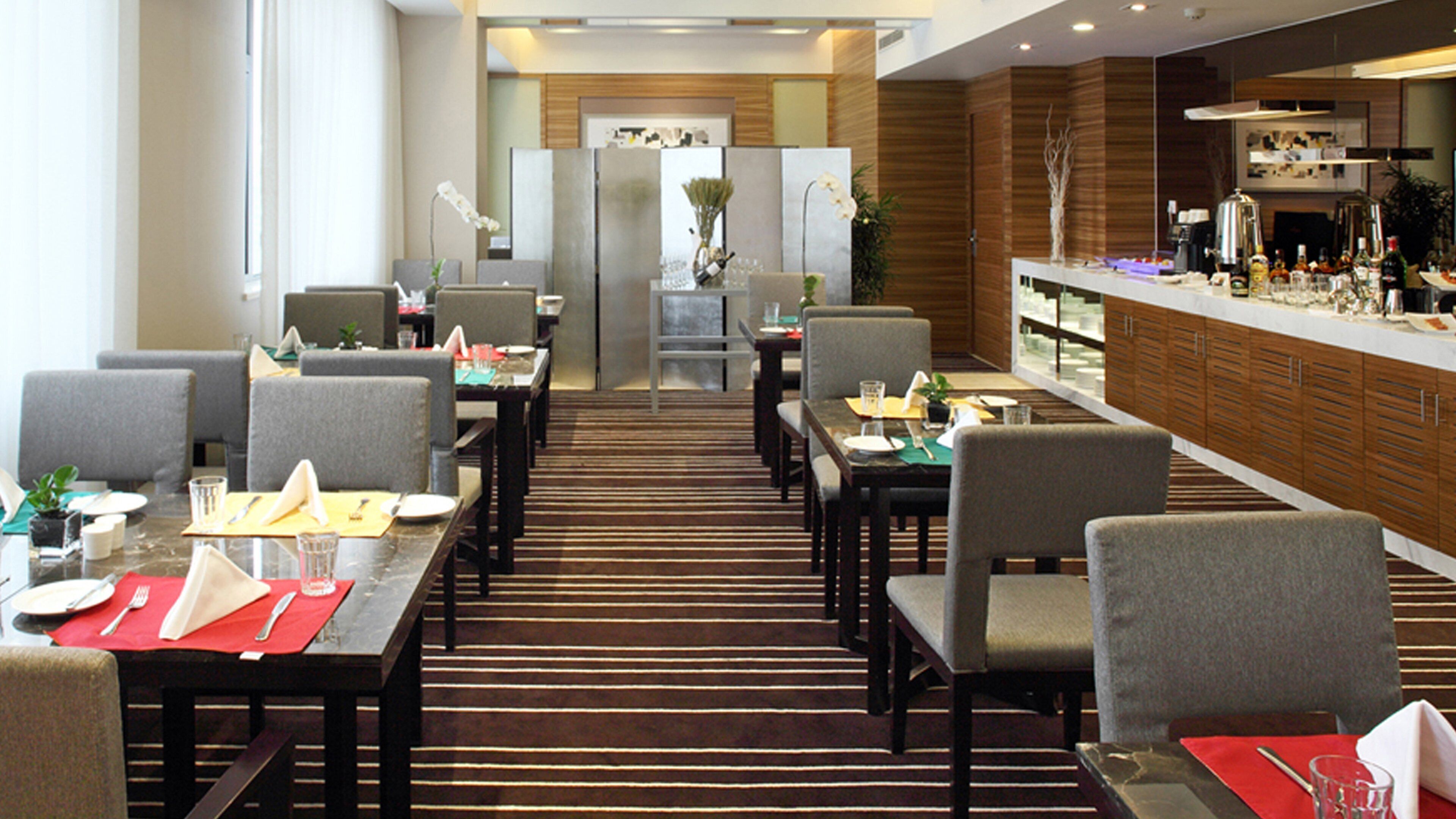 A hotel dining area with multiple tables and chairs, a kitchen area with a bar, and a reception desk in the background.