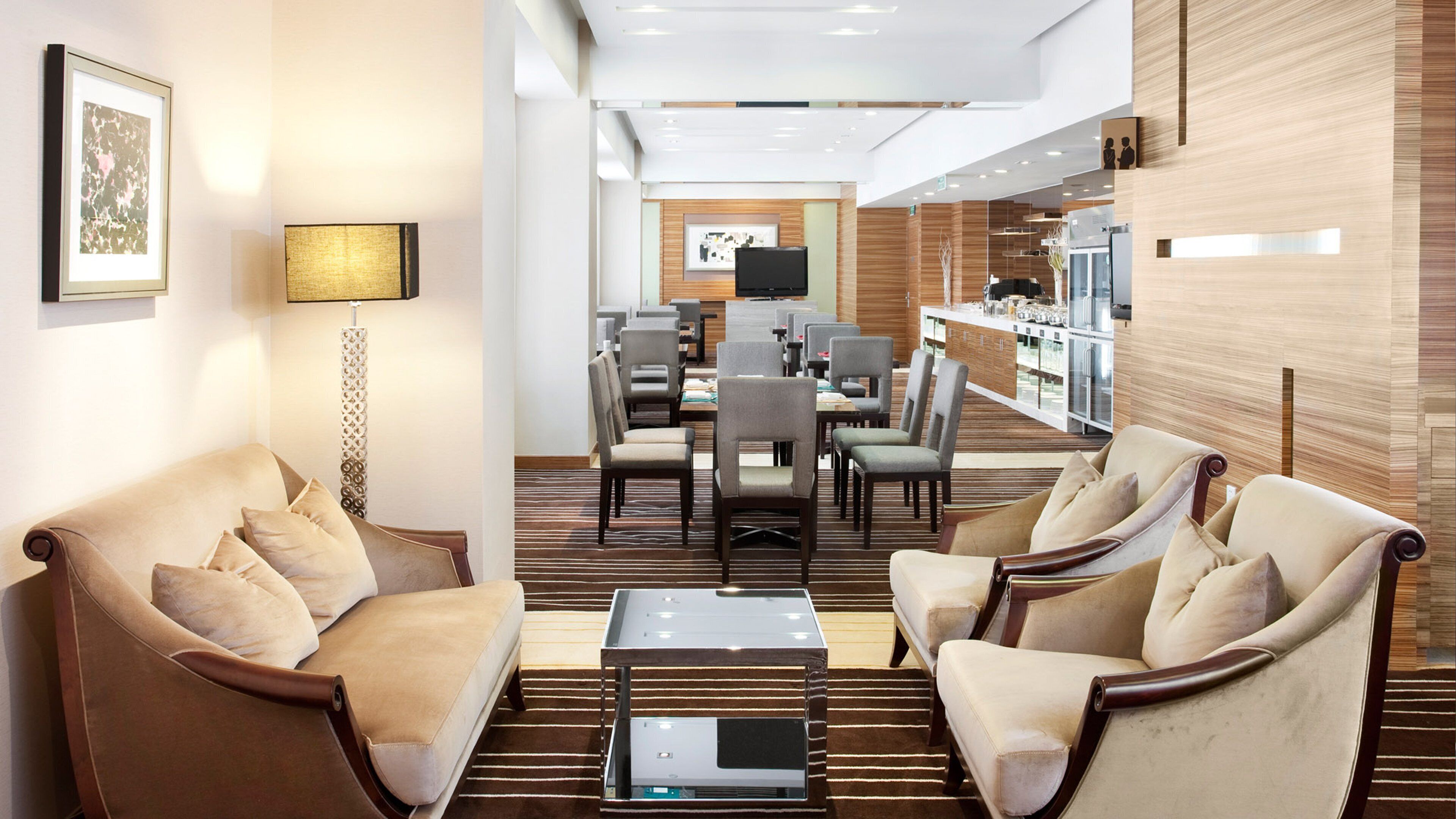 Eine moderne Hotelhalle mit Sitzgelegenheiten, darunter zwei Sofas und ein Couchtisch, ein Essbereich mit Tischen und Stühlen sowie eine Küchenzeile mit Arbeitsplatte und Geräten.