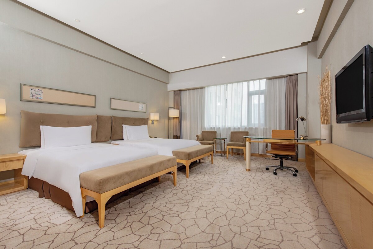 A hotel room with two beds, a desk, a chair, a TV, and a window with curtains.