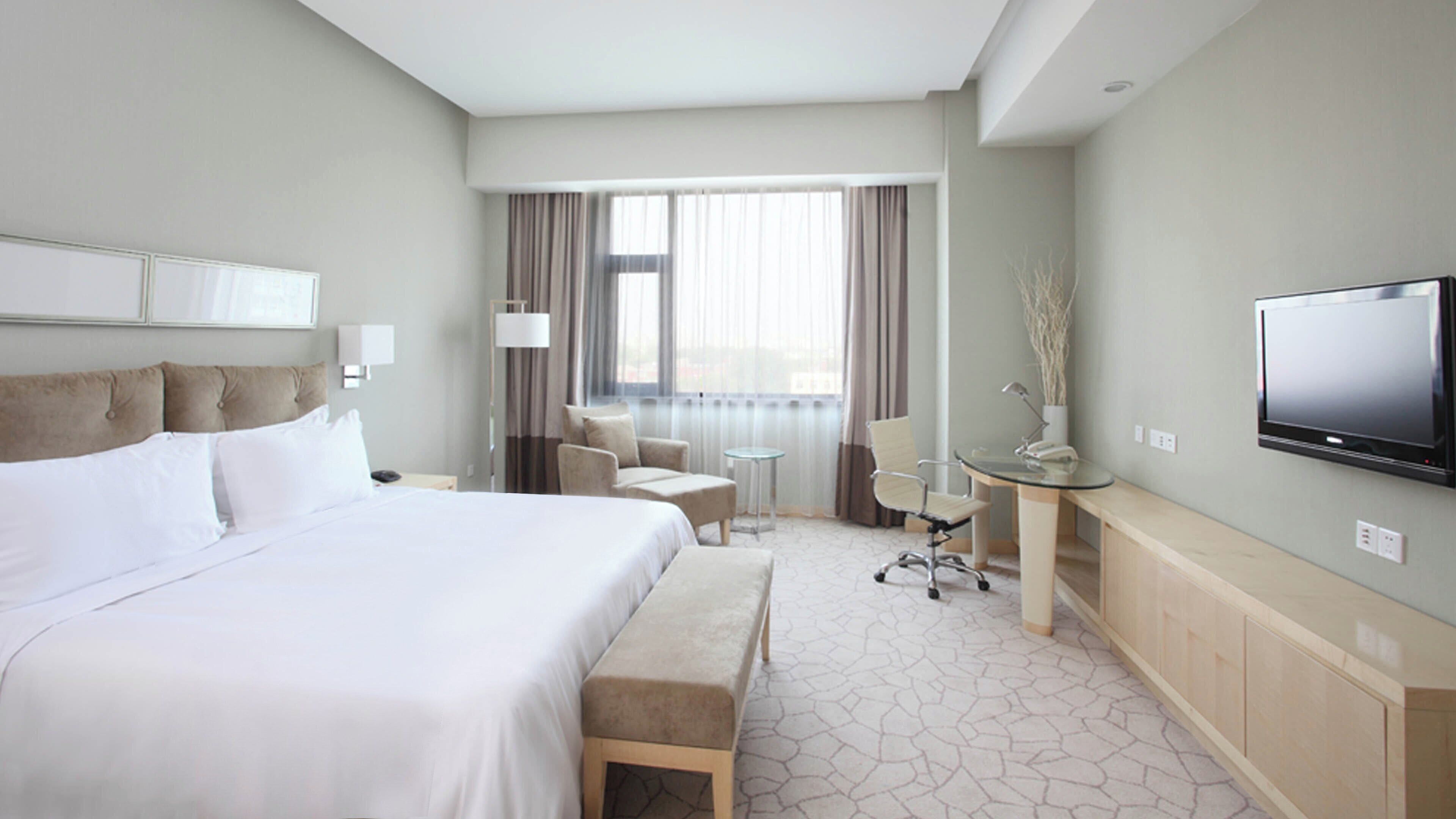 A hotel room with a large bed, a desk with a chair, a television, and a view of the city through the window.