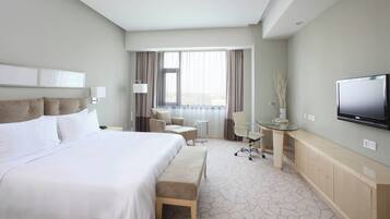 A hotel room with a large bed, a desk with a chair, a television, and a view of the city through the window.