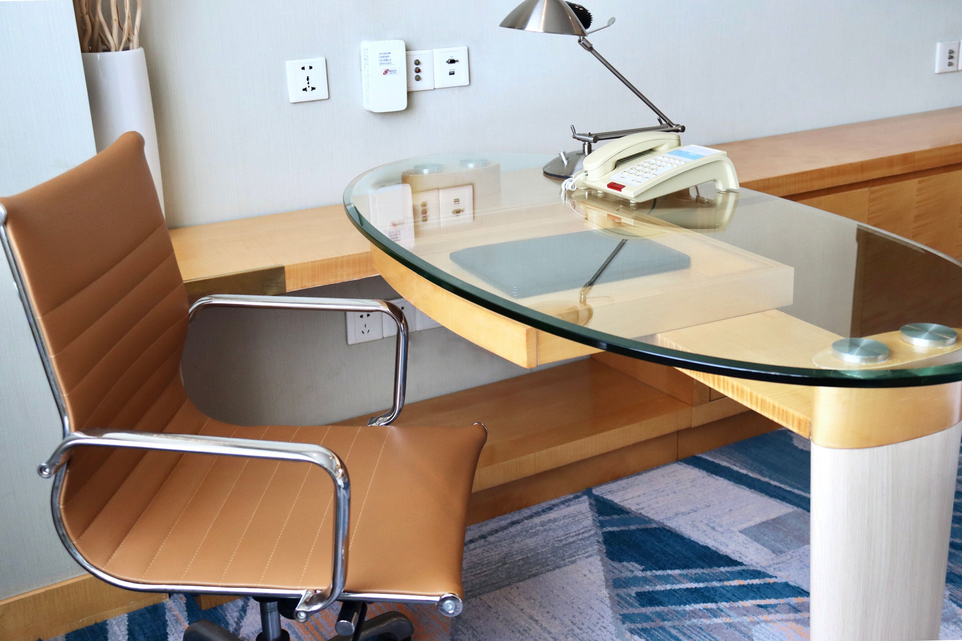 A modern office chair with a high backrest and armrests, a glass-top desk with a telephone, and a lamp.