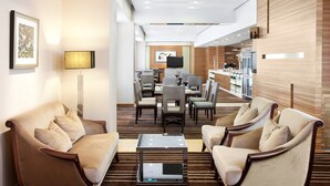 Um lobby de hotel moderno com área de estar com sofás e poltronas, área de refeições com mesas e cadeiras, e uma cozinha visível através de um espaço aberto.