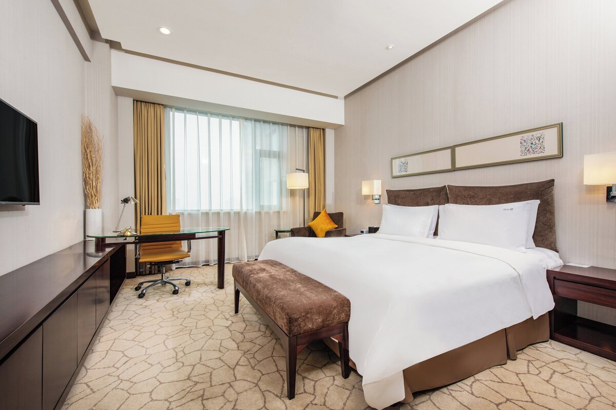 A hotel room with a large bed, a desk with a chair, a bench, a TV, and a window with curtains.