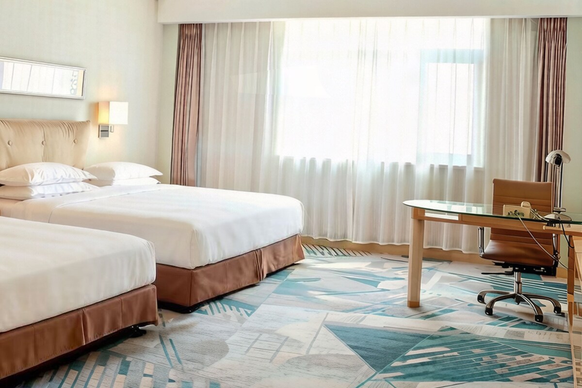 A hotel room with two beds, a desk, a chair, and a large window with curtains.