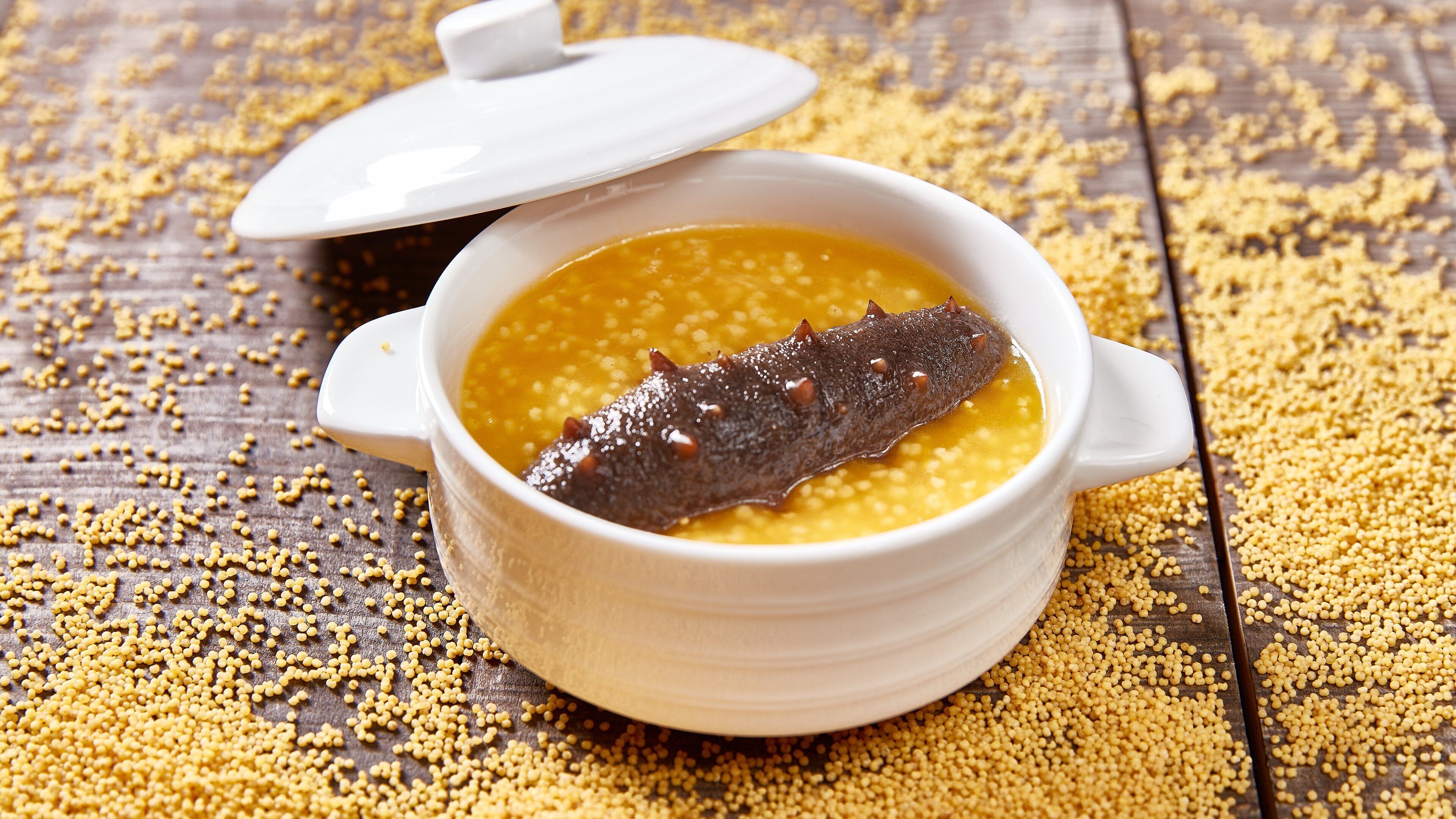 A white ceramic bowl with a lid, containing a yellowish liquid and a dark object, possibly a fish, on a wooden surface with scattered yellow grains.