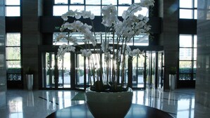 Um vaso grande e de cor escura com orquídeas brancas sobre uma superfície reflexiva num hall amplo, com grandes janelas e vista para o exterior.