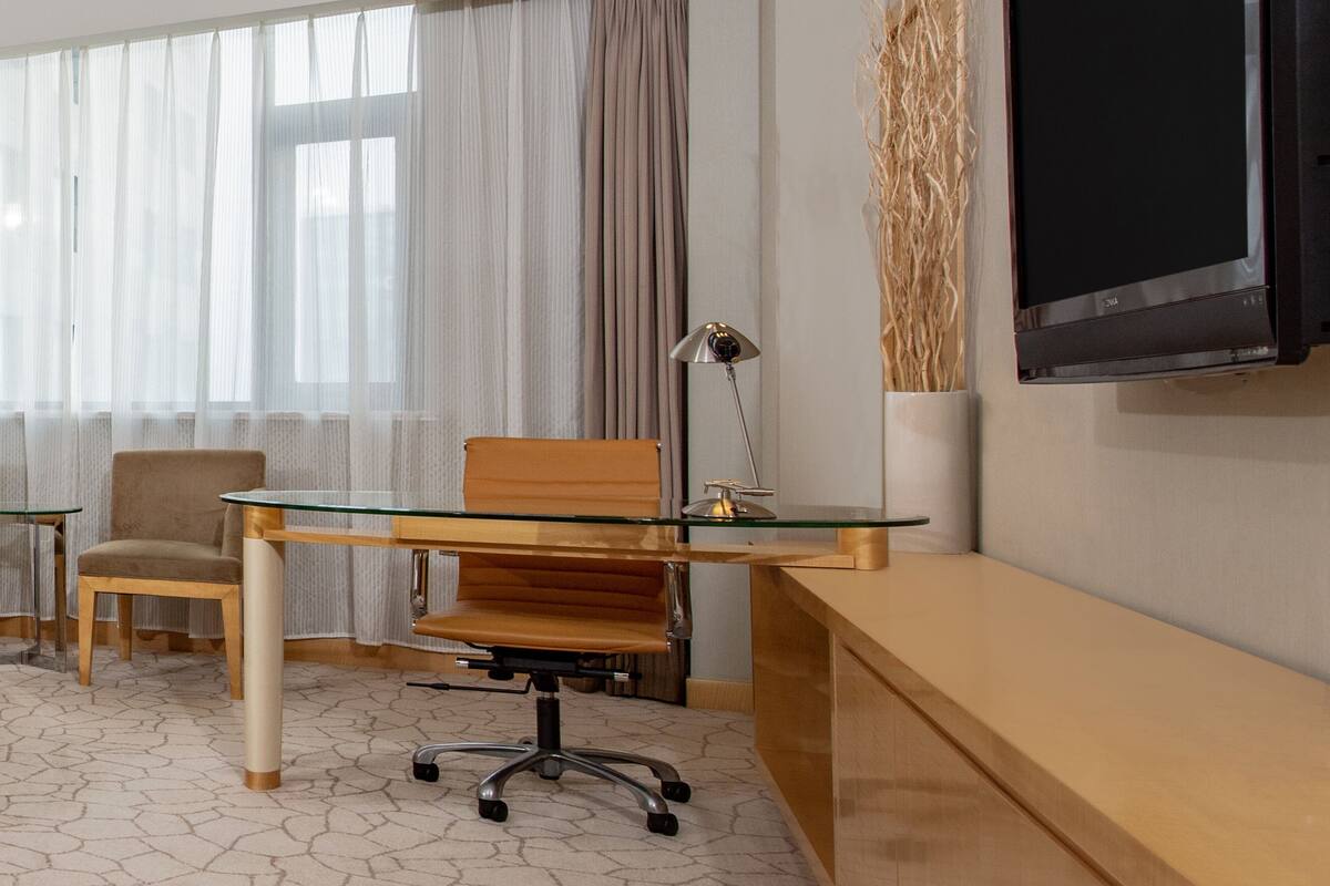 A hotel room with a glass-top desk, a brown leather chair, a lamp, and a flat-screen TV mounted on the wall.