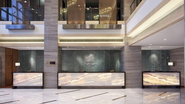 Um lobby de hotel moderno com balcões de recepção em mármore, teto alto e vista do céu através de grandes janelas.