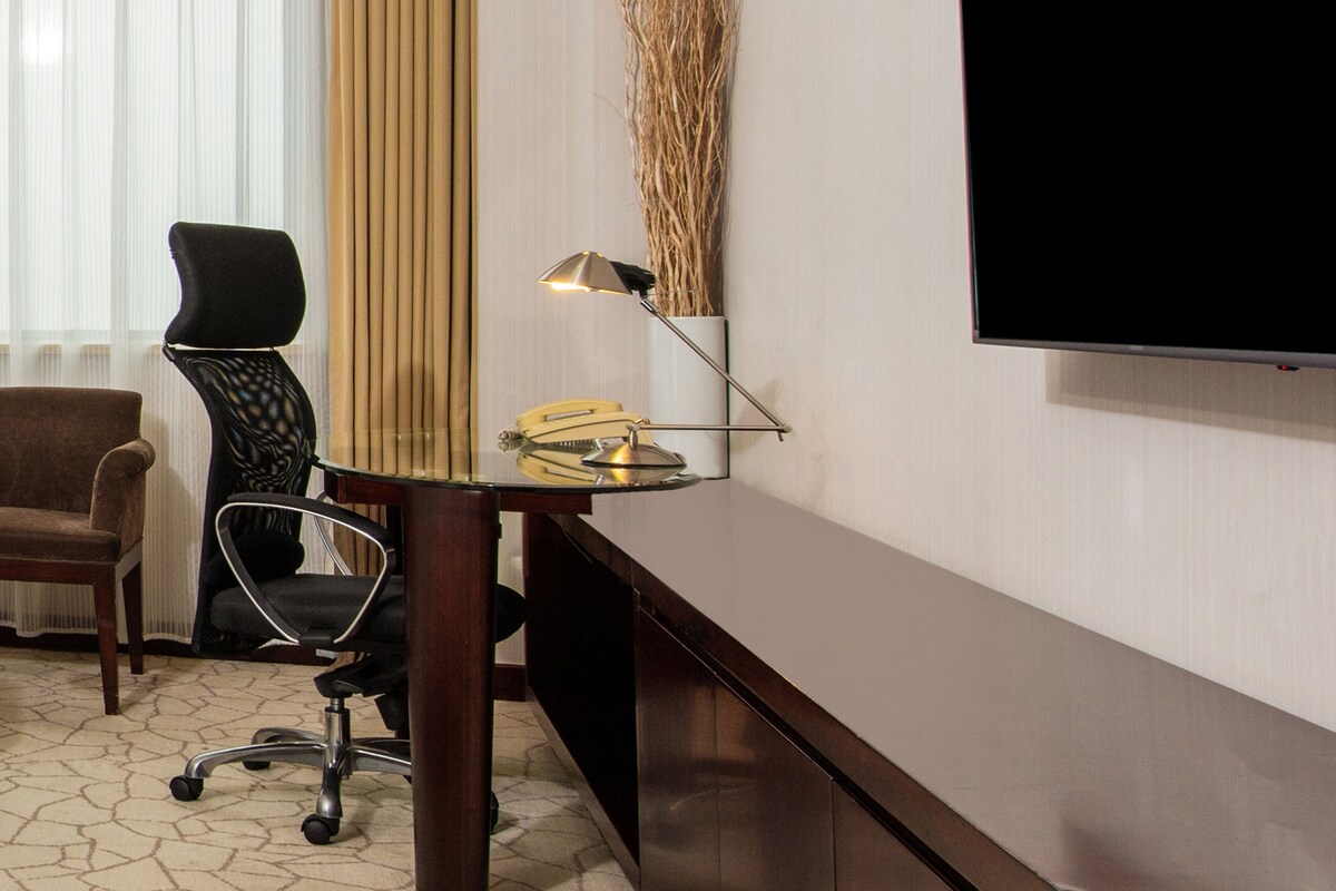 A hotel room with a desk, two chairs, a lamp, a television, and a window with curtains.