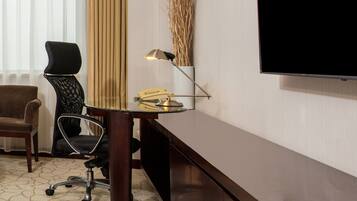Une chambre d’hôtel équipée d’un bureau, de deux chaises, d’une lampe, d’une télévision et d’une fenêtre avec des rideaux.