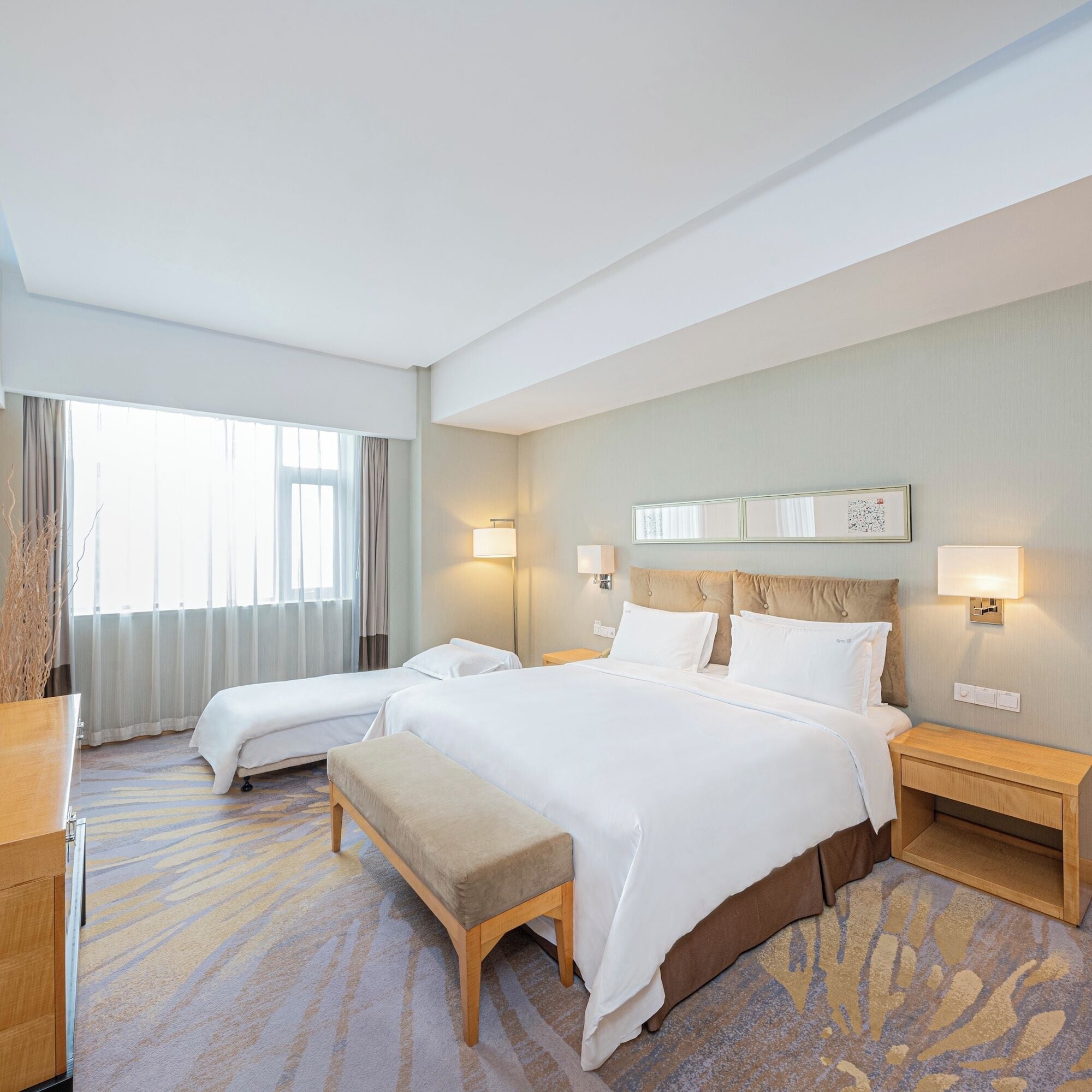 A hotel room with two beds, a bench, a bedside table, and a large window with curtains.