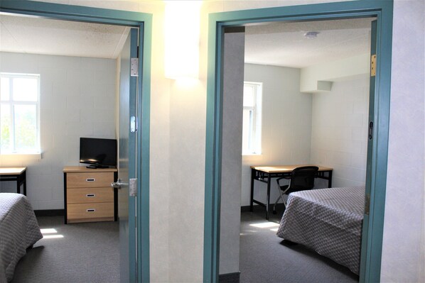 A dormitory room with two beds, a desk, a chair, and a TV.