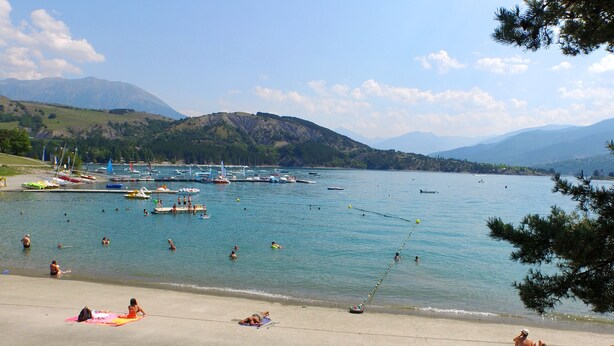 Une plage où des gens se prélassent au soleil et se baignent, des bateaux amarrés dans l’eau, et des montagnes en arrière-plan.