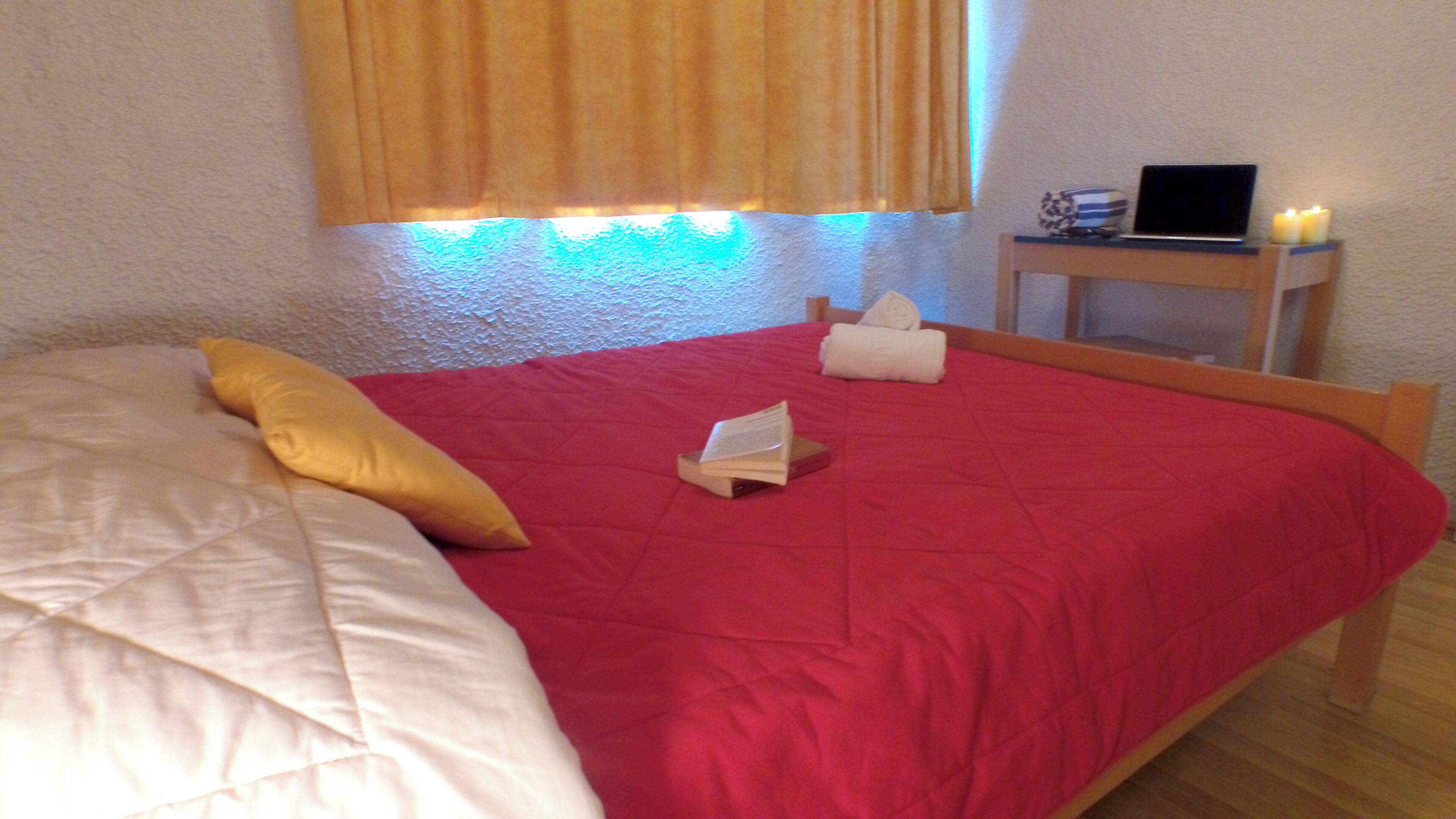 A bed with a red bedspread, a white pillow, and a yellow pillow. There is a small table with a laptop and a candle.