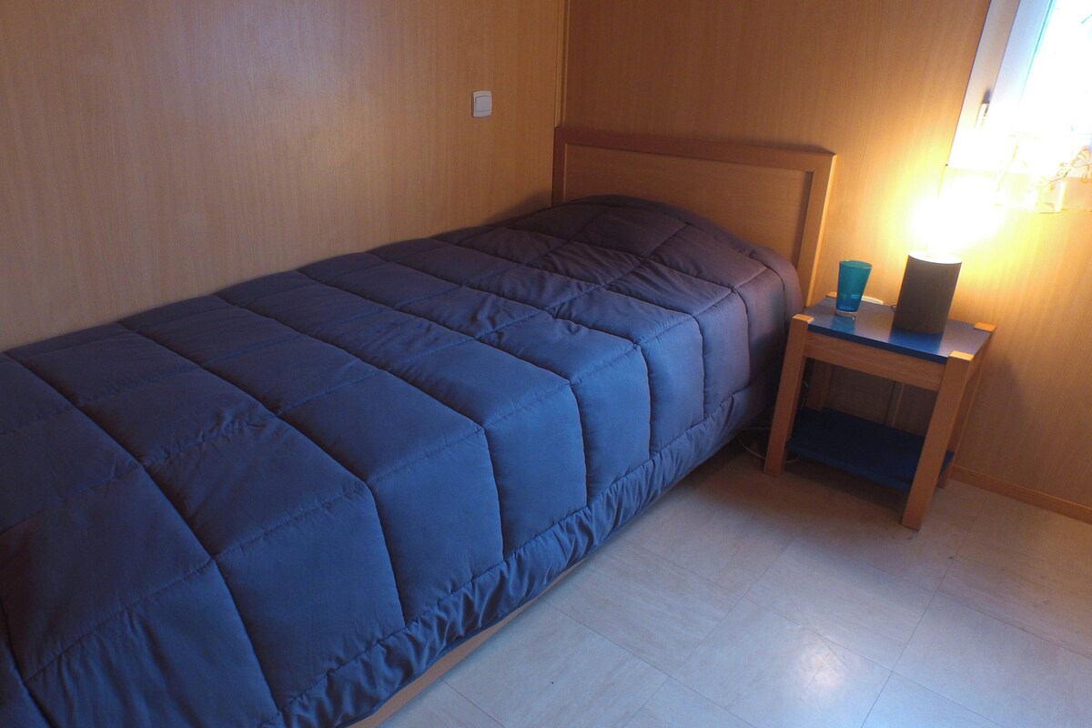 Un lit simple avec une couette bleue, une tête de lit en bois, une table de chevet avec une lampe et une prise électrique au mur.
