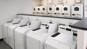 Una lavanderia con diverse lavatrici e asciugatrici bianche ad accesso frontale, un piano di lavoro grigio e una parete rivestita di piastrelle bianche.