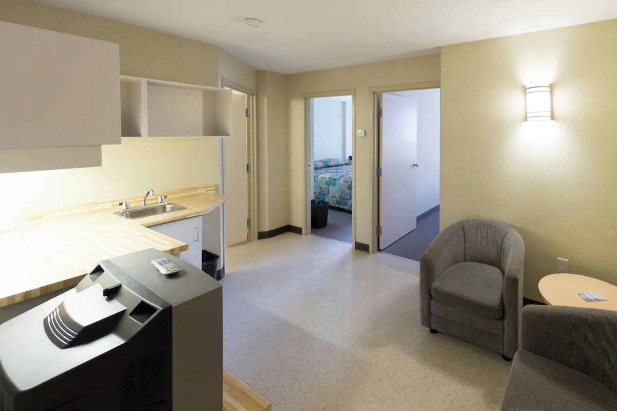 A hotel room with a kitchenette, a grey armchair, a small round table, and a bed with a colorful bedspread.