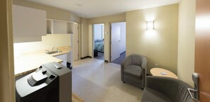 Habitación de hotel con kitchenette, un sillón gris, una mesita redonda y una cama con colcha colorida.
