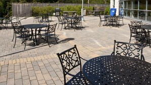 Un'area patio esterna con diversi tavoli e sedie, un barbecue e un edificio con ampie finestre sullo sfondo.