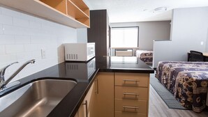 A compact kitchenette with a sink, microwave, and cabinets, a bed with patterned bedding, and a window providing natural light. - Residence & Conference Centre - Kitchener Waterloo (Kitchener)