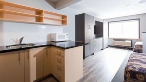 A modern kitchen with a microwave, sink, and wooden cabinets. There is a window providing natural light and a bed with patterned bedding in the adjacent room. - Residence & Conference Centre - Kitchener Waterloo (Kitchener)