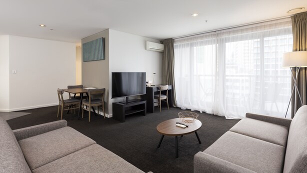 A modern hotel room with a flat-screen TV, a dining area with a table and chairs, a sofa, a coffee table, and large windows with curtains. - Oaks Auckland Hotel (Auckland)