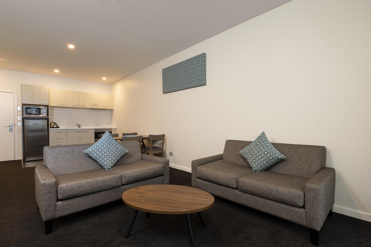A modern living room with two grey sofas, a wooden coffee table, and a kitchen in the background.