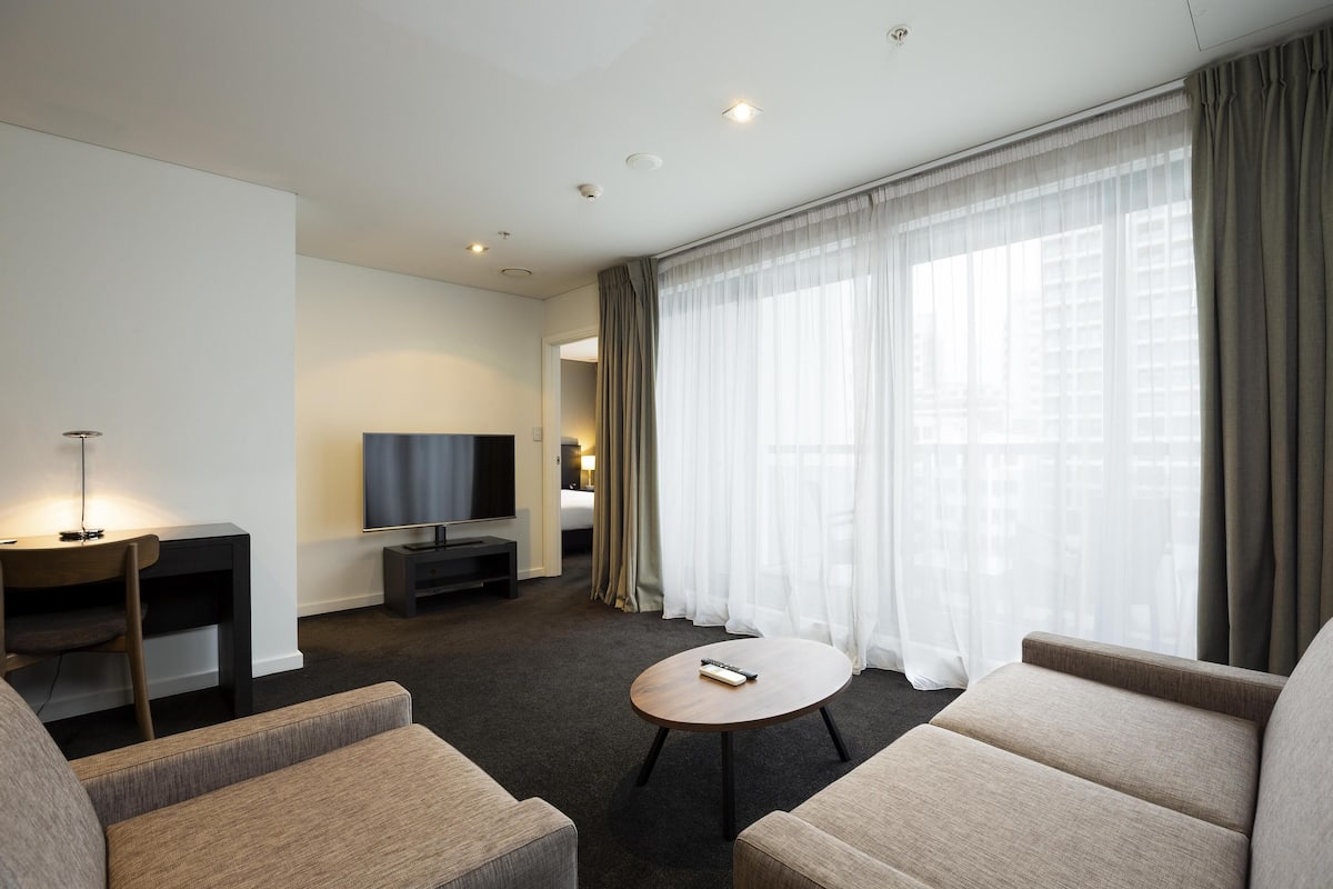 A hotel room with a sofa, a small table, a TV, and a large window with curtains.