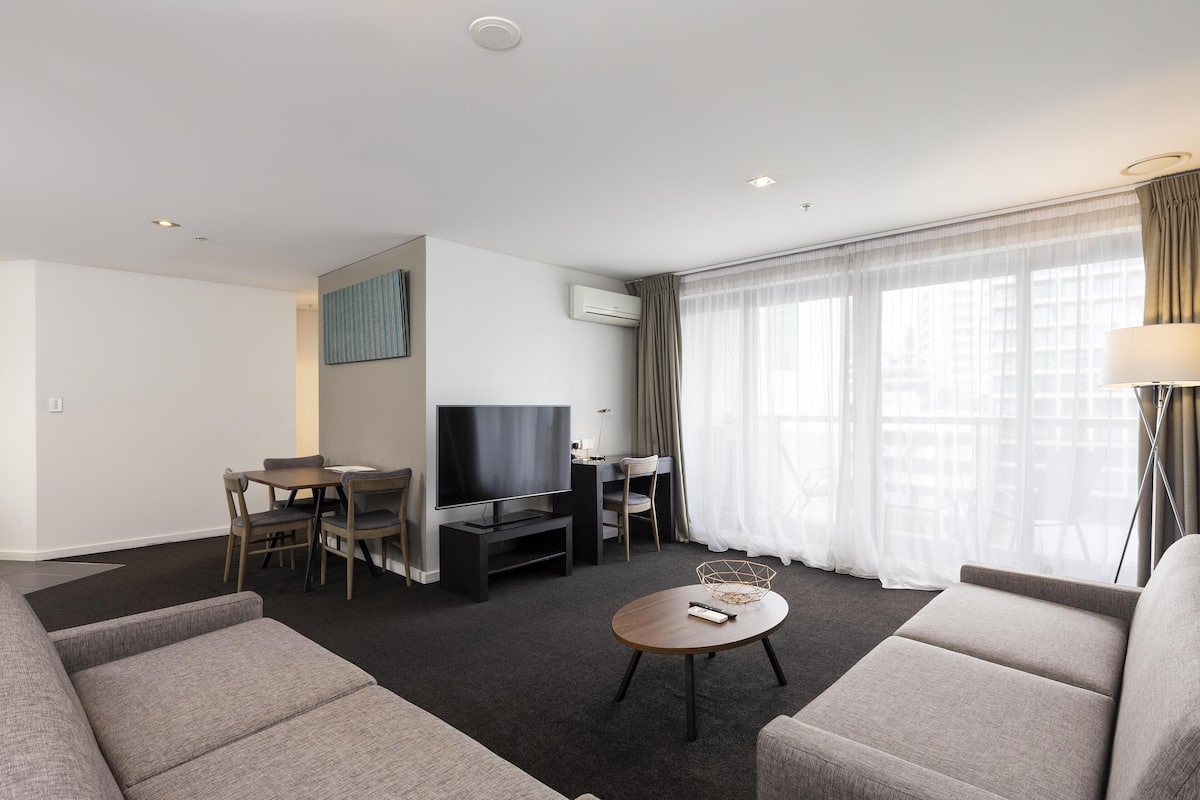 A modern hotel room with a sofa, a dining table, a TV, and a large window with curtains.