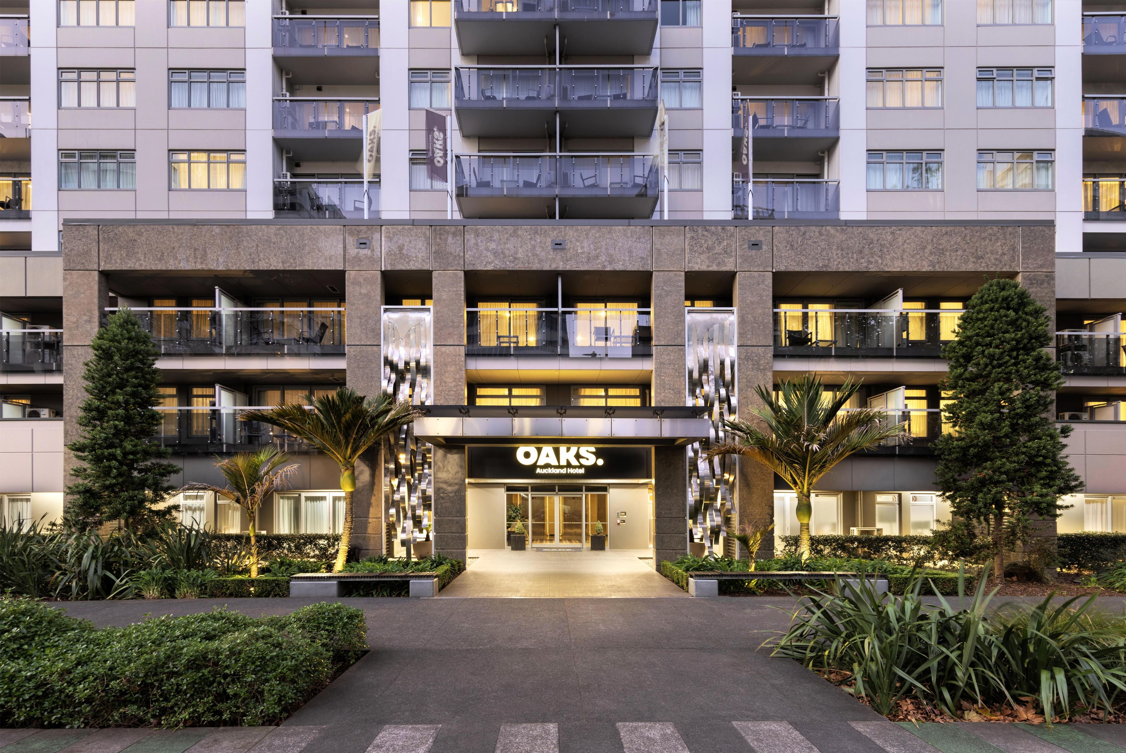 Un hotel de varios pisos con una entrada central, balcones y plantas en el exterior.