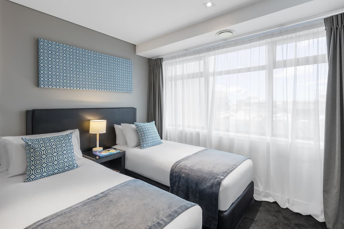 A hotel room with two beds, a nightstand, a lamp, a large window with sheer curtains, and a geometric-patterned wall art piece.