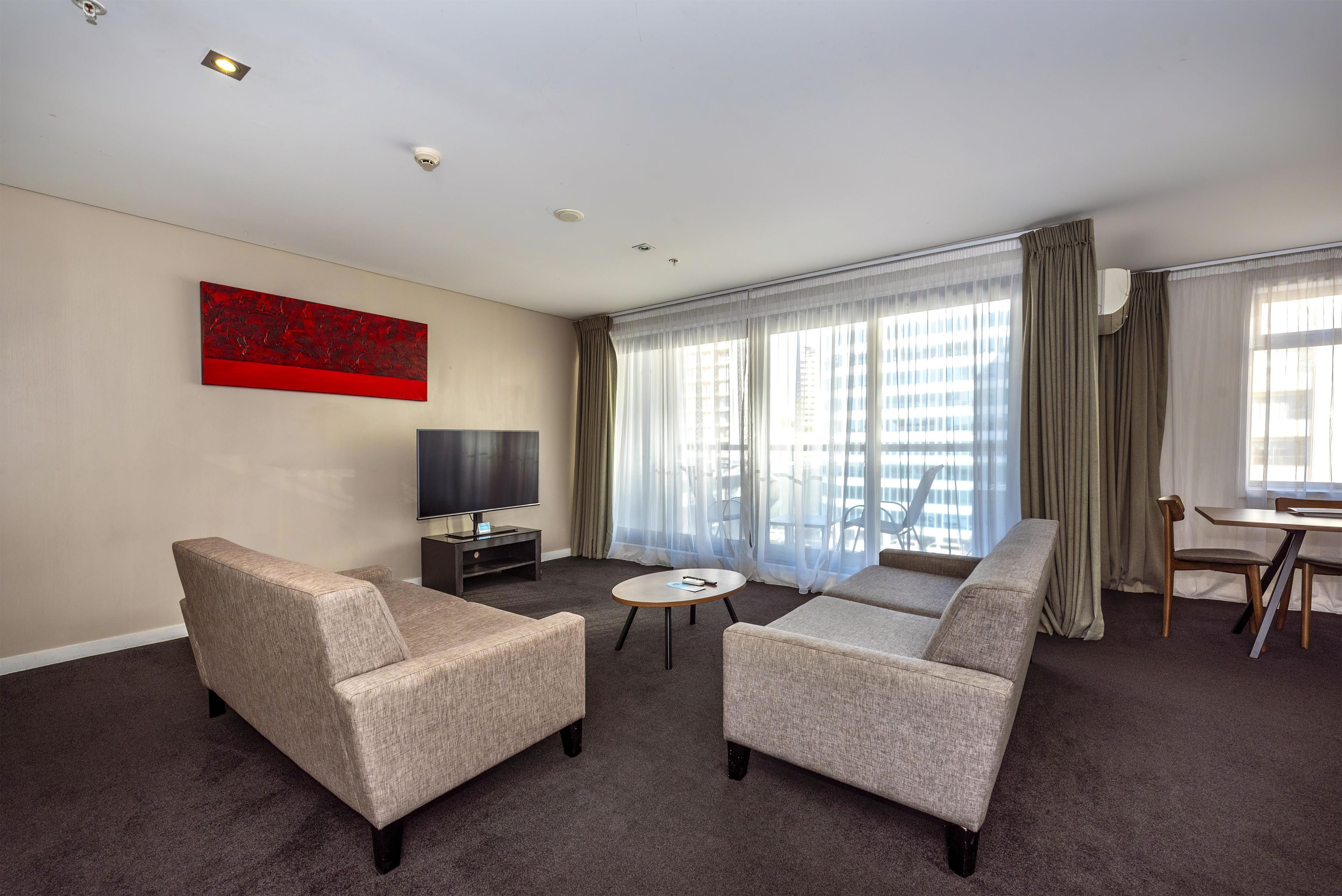 Une chambre d’hôtel avec deux fauteuils, une petite table, un téléviseur et une grande fenêtre avec des rideaux.