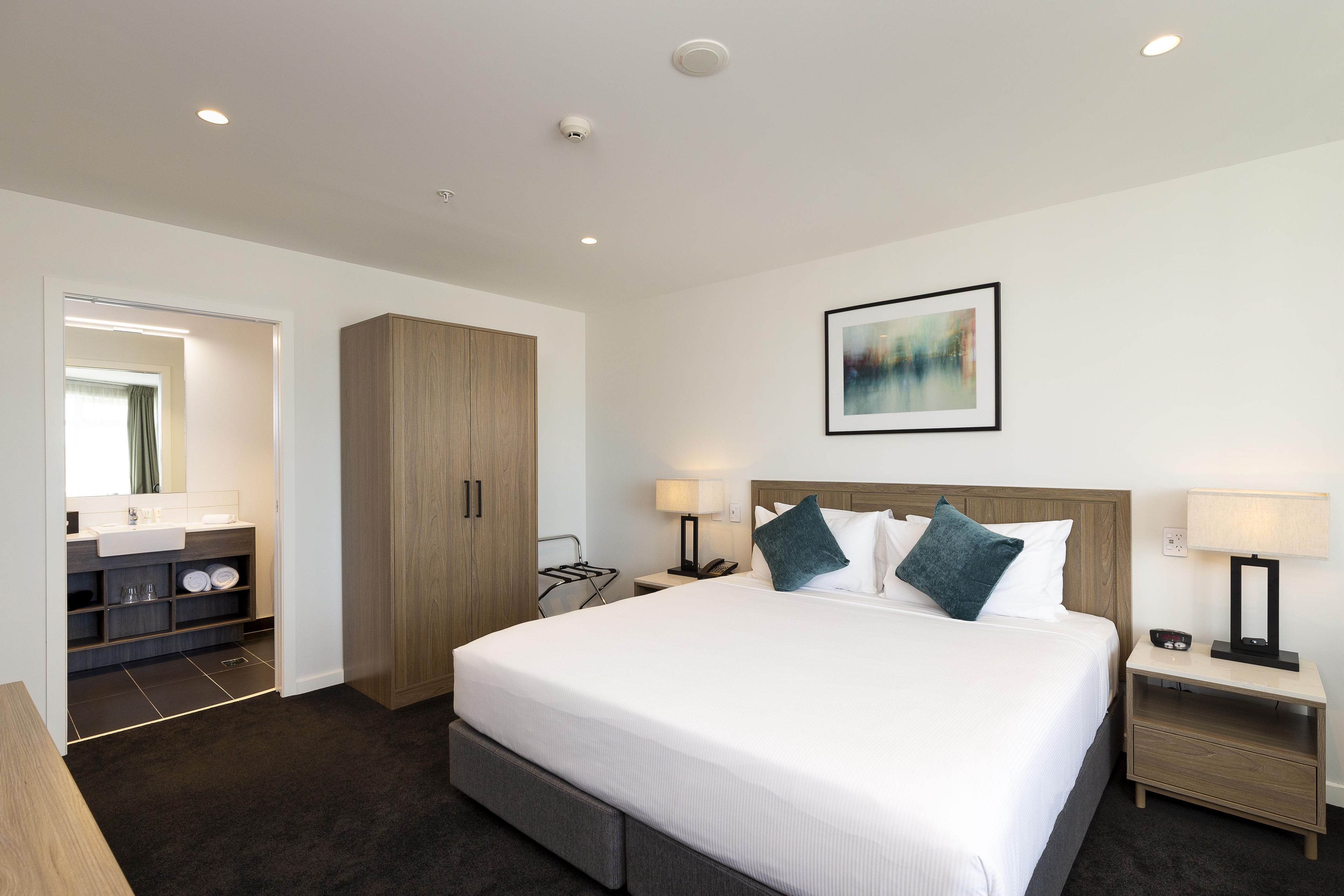 Une chambre d’hôtel avec un grand lit, des lampes de chevet, une table de chevet, une armoire en bois, une salle de bain avec un lavabo et un miroir, et un tableau abstrait encadré accroché au mur.