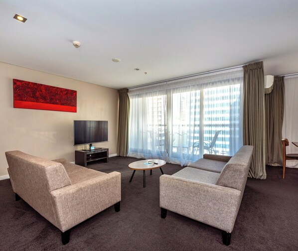 Una habitación de hotel con sofá, sillones, televisión y un ventanal con cortinas.