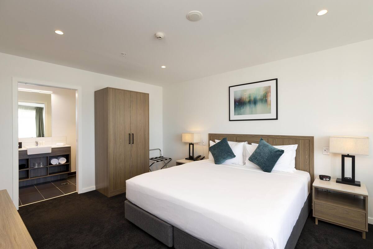 A modern hotel room with a large bed, a wooden wardrobe, a bathroom with a sink and mirror, and a painting on the wall.