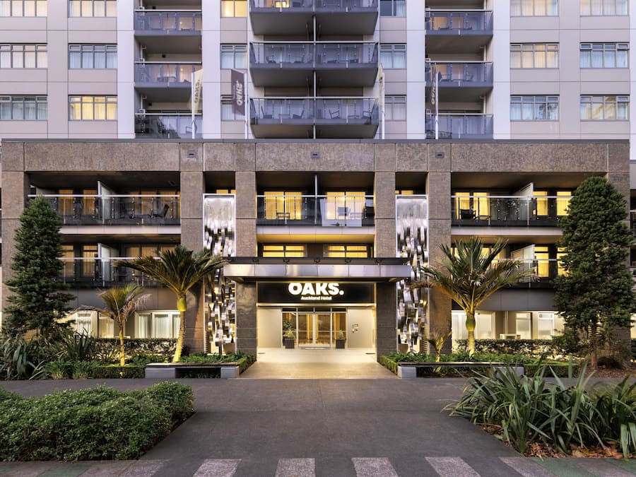 Un hotel de varios pisos con una entrada central, balcones y plantas en el exterior.