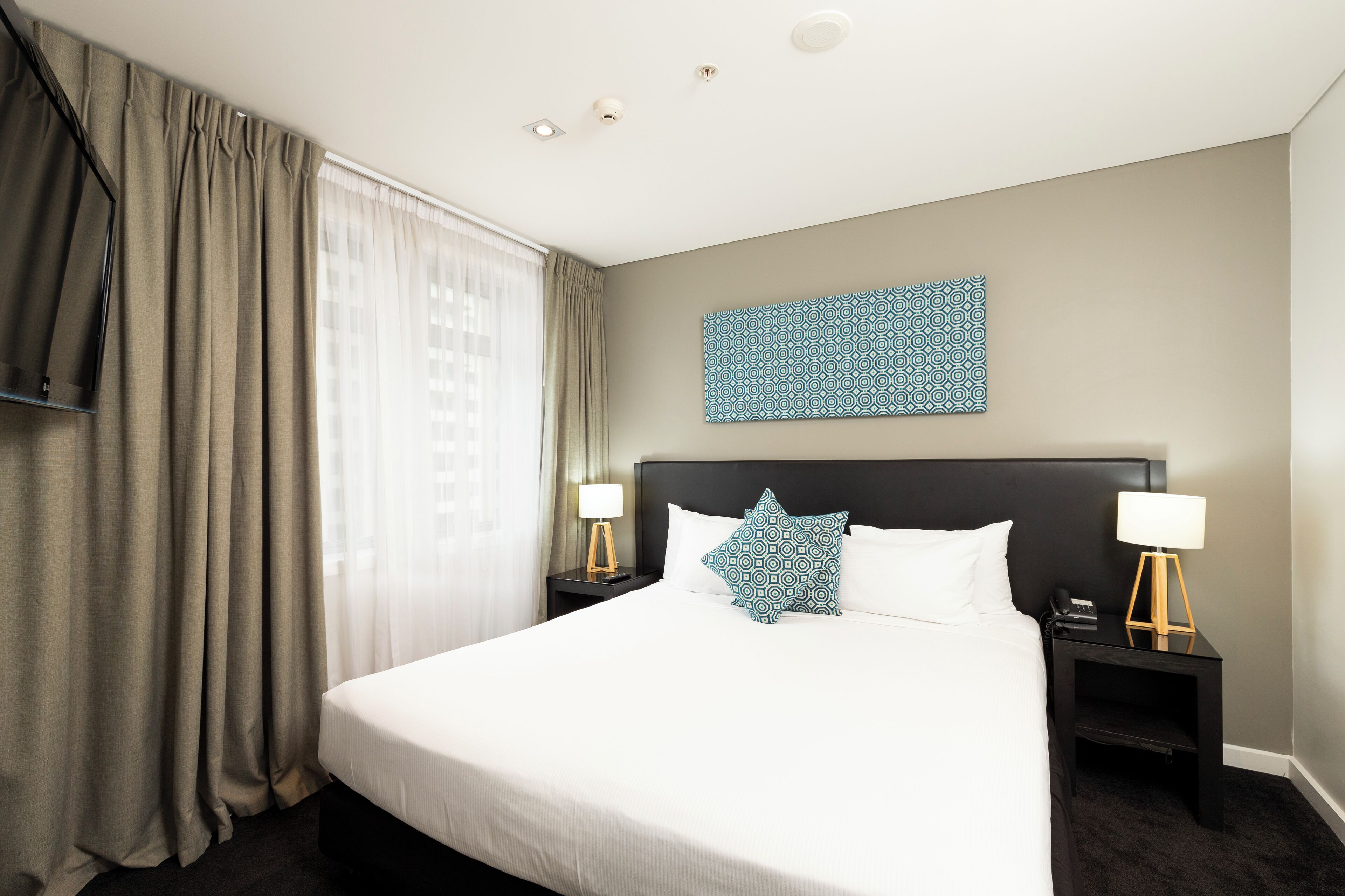 Een hotelkamer met een groot bed, twee nachtkastjes met lampen, een tv aan de muur en een kunstwerk met patroon boven het bed.