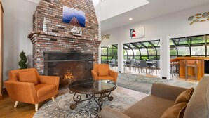 Un espacio moderno con zona de estar, chimenea de ladrillo, sillones naranja, una mesa de centro de vidrio y vistas al exterior a través de amplias ventanas.