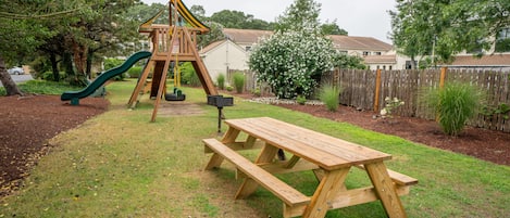A wooden picnic table and a playground with a slide and climbing structure in a backyard.