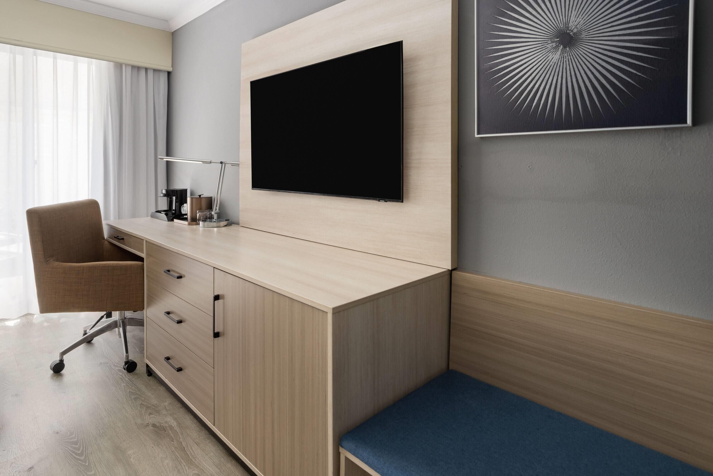 A modern hotel room with a large flat-screen TV mounted on the wall, a desk with a chair, a small bedside table, and a blue cushioned bench.