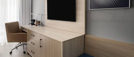 A modern hotel room with a large flat-screen TV mounted on the wall, a desk with a chair, a small bedside table, and a blue cushioned bench.
