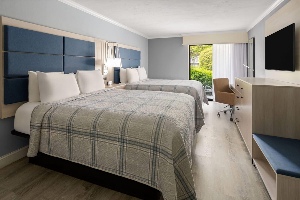 Une chambre d’hôtel avec deux lits, un bureau, une chaise, une télévision et une fenêtre donnant sur un espace verdoyant.