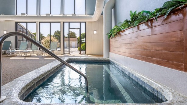 Una piscina interior con vista despejada al exterior, una pared de madera con vegetación y una zona de estar con sillas.