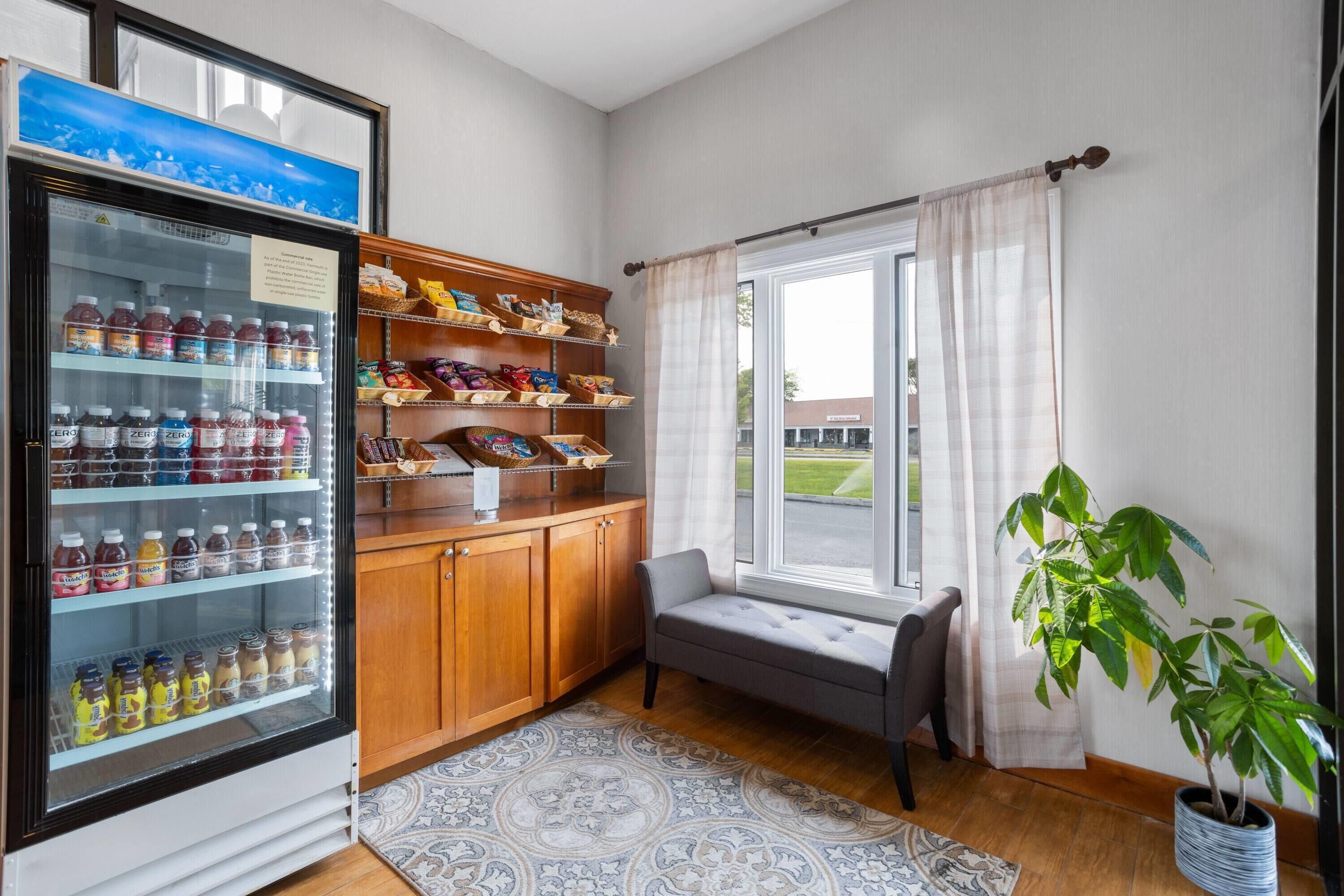 A room with a refrigerator stocked with beverages, wooden shelves with snacks, a bench, a potted plant, and a window with curtains.