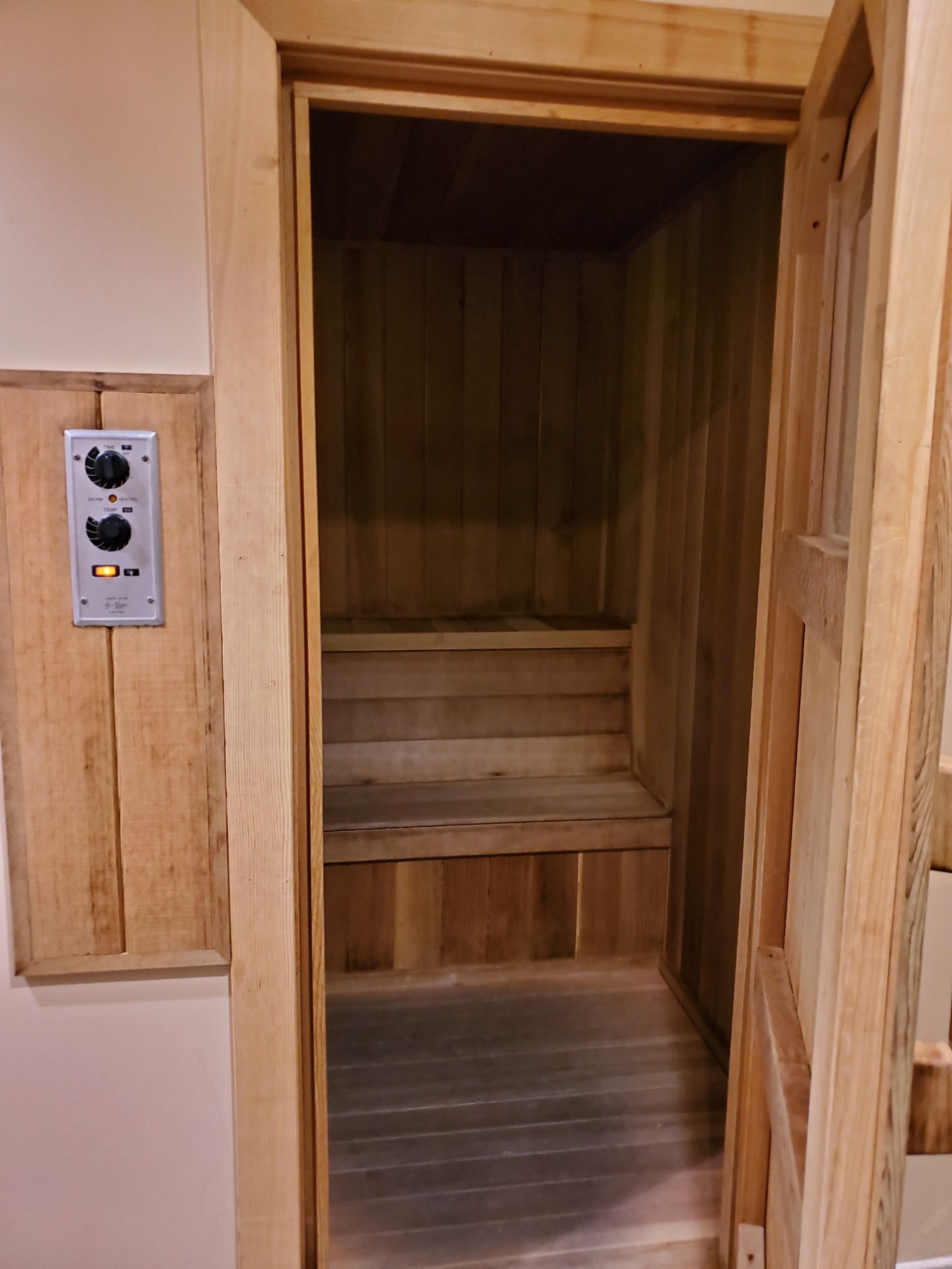 A wooden sauna with a control panel on the wall and a set of wooden steps leading into the sauna.